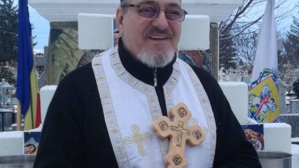 Preot ortodox cu o cruce in mana in interiorul unui cimitir.